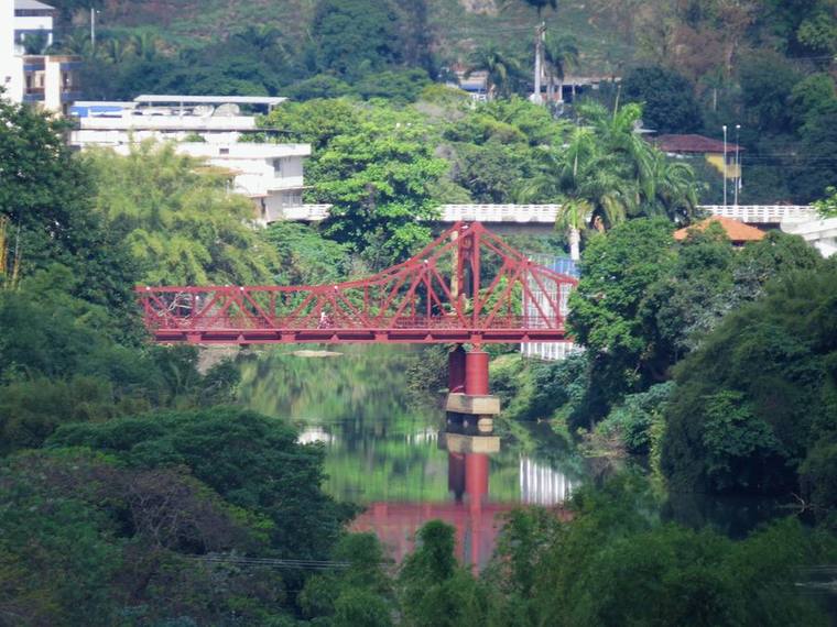 The Ponte Metálica, a red steel truss bridge crossing the Rio Pomba between wooded banks.